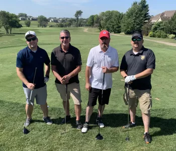 Four men stand ready to golf with drivers on a golf course next to houses and trees, one sporting a cigar in his mouth