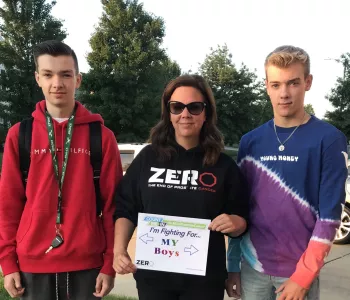 A mother sporting a Zero prostate cancer hoodie holding a sign that says "I'm fighting for my boys"