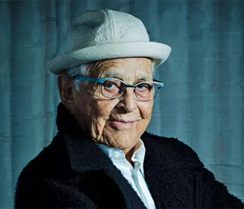 TV writer and personality, Norman Lear, wearing a white hat and dark jacket while sitting in front of a gloomy blue background