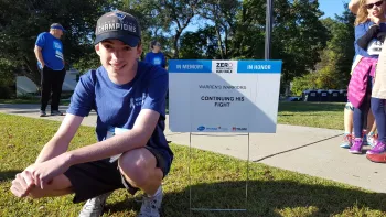 A younger teen aged boy kneeling next to a yard sign  at an event that says Warrens Warriors