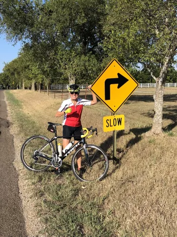 Jan C Johnson on a bike by a Slow Right Corner sign
