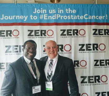 Mike Crosby and Chas Rodgers stand in front of a journey to end prostate cancer step and repeat