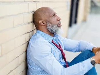 Black man sitting on the ground against a brick wall looking distressed