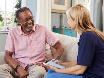 An older Black man sitting on a couch with a woman who is holding a clipboard