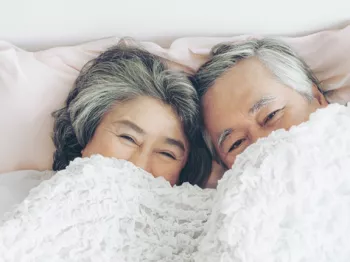 Senior asian couple laying in bed with the sheets covering half their faces.