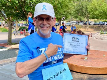 A senior man, Bob Jacobs, at a ZERO Run/Walk wearing a white hat with a blue ribbon, a light blue ZERO Run/Walk t-shirt with a bib that says "Bob's Baby Blues," and holding an award for Top Rookie Team of the year