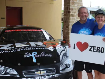 Joni and Husband holding up I Heart ZERO sign by a car with a large ZERO logo on it