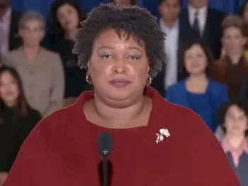 Stacy Abrams speaking at a microphone wearing a classic red sweater
