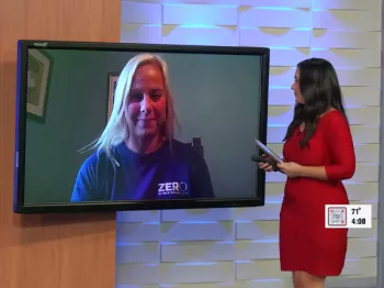  A woman wearing a ZERO shirt appears on a tv screen that a news anchor wearing a red dress is standing next to