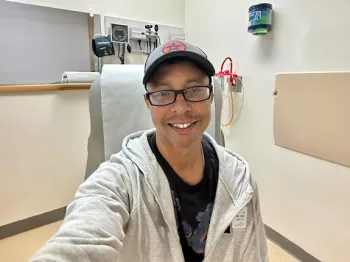 A man wearing a beige zip-up hoodie over a black tshirt, glasses, and a black hat, taking a selfie from a hospital room.