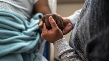 Two black people holding hands; one of them is in a hospital bed