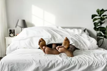 The feet of two Black people sticking out from under the covers of a large bed