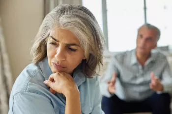 An older Hispanic woman with gray hair with her hand under her chin as she is turned away from a man sitting behind her in an aggressive stance as if they are arguing