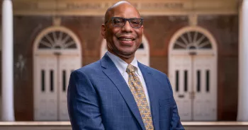 Brian Bragg, Chief Mission Officer, a Black man in a blue suit and yellow checkered tie standing in front of a brick building