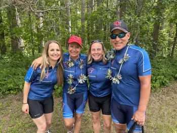 A group of three women and one man all wearing cycling jerseys that says "Miles for Money" stand for a group photo