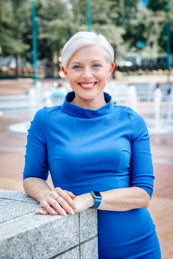 Blonde woman with short hair wearing a blue dress standing in front of a fountain