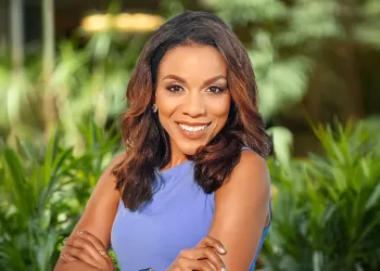 A Black woman in a purple top standing with her arms crossed and smiling in front of green foliage