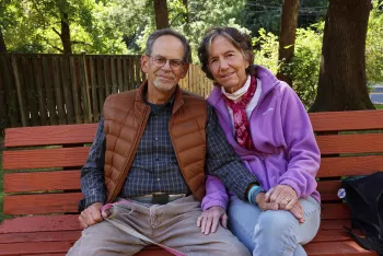 A couple sits together on a red-painted wooden bench in a backyard or park setting. One person wears glasses, a brown puffy vest over a plaid shirt, and khaki pants. The other wears a bright purple fleece jacket with a red bandana and light-colored jeans. They are both smiling warmly at the camera with trees and a wooden fence visible in the background on a sunny day.