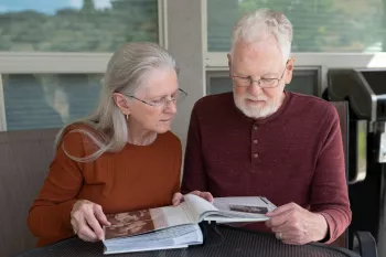 A senior couple sits together at a table looking at what appears to be a photo album. The woman has long silver hair and wears a rust-colored sweater, while the man wears a burgundy henley shirt and has glasses. They are both engaged in viewing the album's contents in what appears to be a bright setting.