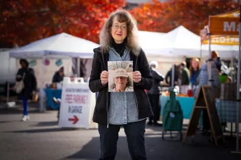 At an outdoor event with white tents and vibrant red autumn trees in the background, a woman with long wavy gray hair and glasses stands wearing a black jacket over a denim shirt. She holds up a photo labeled "Heroes" while standing in what appears to be a market or fair setting. White event tents, vendor displays, and other attendees can be seen behind her.