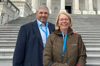 Two individuals wearing blue lanyards stand smiling on the steps of a grand building with white columns and a 'Members Only' sign in the background. The man wears a dark suit, while the woman wears a brown Columbia jacket and glasses.