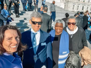 A group of people smiling on the steps of a large building, with some wearing blue scarves that say "ZERO Prostate Cancer." The setting appears to be a formal gathering or event.