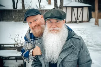 Two men wearing flat caps pose together on a snowy winter day. Both have full beards - the man in front has a long, flowing white beard and wears a gray coat, while the man behind him has a shorter reddish beard and wears a plaid shirt. They are smiling warmly at the camera and appear to be in a backyard setting with trees and a fence visible in the background.