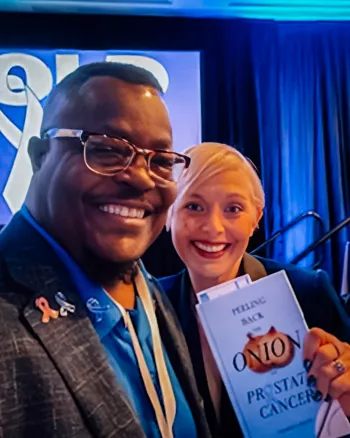 Darnell and Courtney smile for a selfie at an indoor prostate cancer awareness event. Darnell wears glasses and a blue suit jacket with an awareness ribbon pin, while Courtney holds up a book titled “Peeling Back the Onion on Prostate Cancer.” A large blue awareness ribbon is visible in the background.