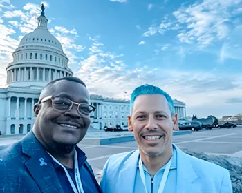 Darnell and Leo pose in front of the U.S. Capitol building under a bright blue sky. Darnell wears a blue suit and glasses, and Leo stands beside him with bright blue hair and a matching blue jacket, both wearing conference lanyards.