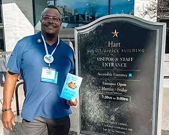 Darnell stands outside the Hart Senate Office Building, smiling and holding the book “Peeling Back the Onion on Prostate Cancer.” He wears a navy blue polo shirt with conference badges hanging from a lanyard around his neck. The building sign is clearly visible beside him.