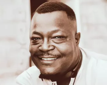 Headshot of a smiling man with close-cropped hair and a neatly trimmed mustache, wearing a light-colored collared shirt. He is looking directly at the camera with a warm and friendly expression. The background is softly blurred, keeping the focus on his face.