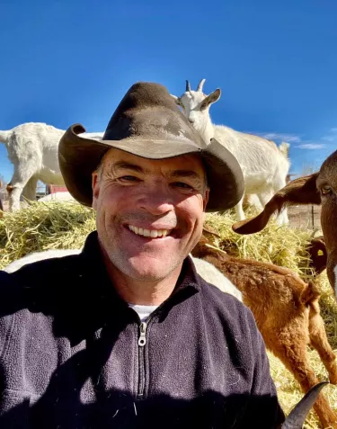Cowboy Max and goat looking at the camera taking a selfie while goats romp on hay piles in the background