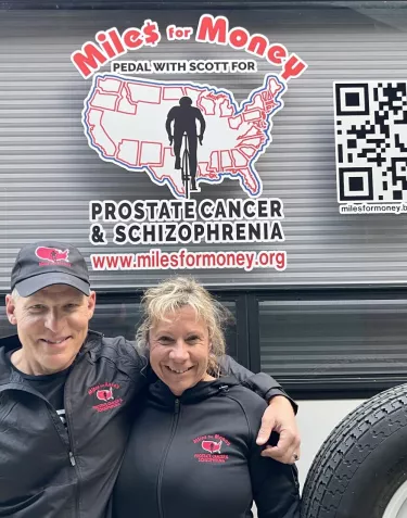 A heterosexual couple dressed up in black sports clothes in front of a truck with a sign Cycling for prostate cancer