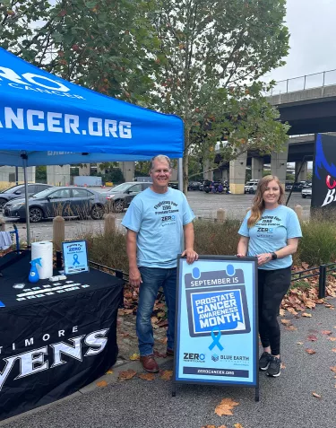 Two volunteers standing outside of the ZERO tent and A-Frame sign promoting Prostate Cancer Awareness Month at the Baltimore Ravens RavensWalk pre-game event in September 2023