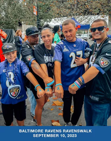 A group of people wearing Baltimore Ravens shirts and jerseys, holding out their wrists to show off prostate cancer awareness bracelets and temporary tattoos on their hands.