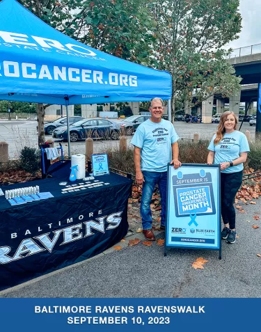 A man and woman wearing prostate cancer awareness t-shirts standing by a sign for ZERO Prostate Cancer Day outside of a ZERO tent with a table underneath that has a Baltimore Ravens tablecloth.
