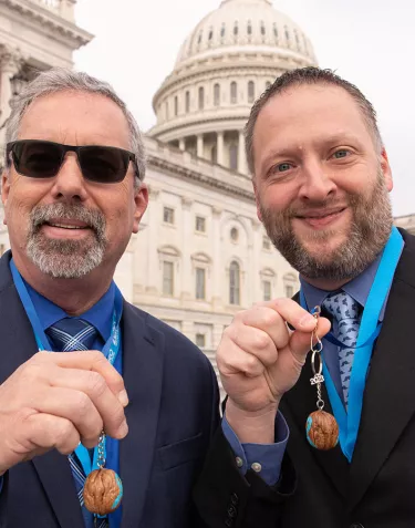 Two men in suits showing a walnut prostate cancer mascot in front of the Capitol