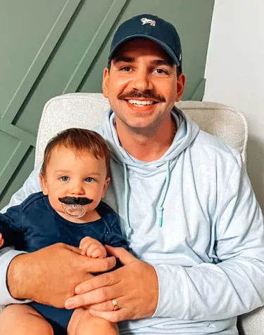 A man with a mustache holding a baby wearing a fake mustache