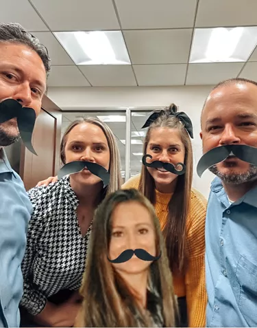 A group of people in an office wearing fake paper mustaches