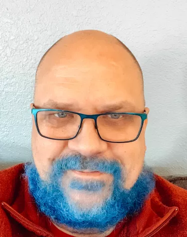 A bald man with glasses with a beard dyed blue