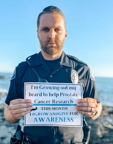A police officer in uniform standing on a beach holding a sign that says he is supporting ZERO through Grow & Give
