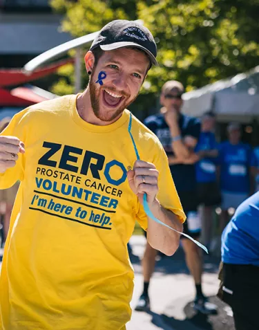 An energetic volunteer wearing a yellow ZERO Prostate Cancer Volunteer t-shirt with "I'm here to help" text smiles broadly at the camera on a sunny day. He has a blue ribbon painted on his face, with other volunteers visible in the background.