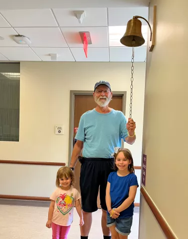 A person in a light blue t-shirt and baseball cap stands with two young girls in a hospital hallway, holding a chain connected to what appears to be a ceremonial bell mounted on the wall. The setting includes white walls, a wooden handrail, and standard hospital ceiling tiles with a red exit sign visible.