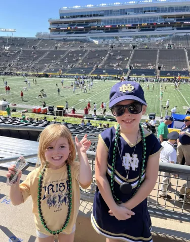 Two young girls at a Notre Dame football stadium, wearing Notre Dame themed clothing and green bead necklaces. One wears a yellow "Irish" t-shirt and the other wears a navy Notre Dame cheerleader-style dress. They're smiling at the camera with the football field and stadium seating visible in the background.