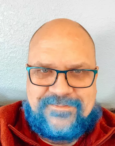 A close-up of a person with a bald head, wearing glasses, and a bright blue beard. They are wearing a red sweater and sitting against a light-colored wall.