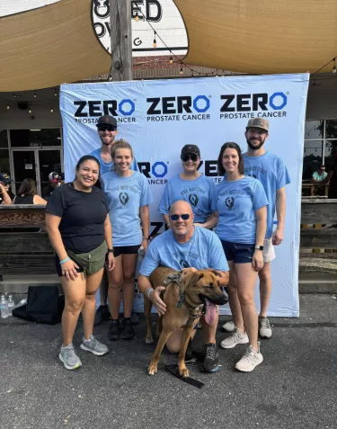 A group photo of volunteers at a ZERO Prostate Cancer event, standing in front of a large blue tent or canopy with "ZEROCANCER.ORG" text. The volunteers are wearing matching light blue t-shirts and are arranged in two rows. 