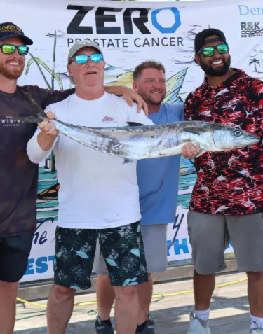 Four men standing together at a fishing event, posing with a large caught fish in front of a ZERO Prostate Cancer branded backdrop with multiple sponsor logos. From left to right: a man in a dark Darwin's Selects shirt and baseball cap, a man in a white t-shirt holding the fish, a man in a light blue shirt, and a man in a red and white patterned long-sleeve shirt. All are wearing sunglasses and smiling.