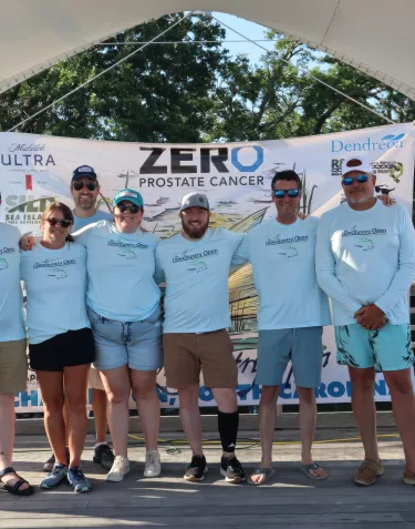 A group photo of people wearing matching light blue ZERO Prostate Cancer t-shirts gathered outdoors for what appears to be a community event or fundraiser. The group includes both adults and children, and they're smiling at the camera with trees visible in the background.