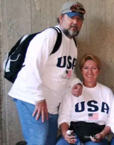 A couple wearing matching USA t-shirts with American flag designs pose casually against a concrete wall. The man wears a baseball cap and carries a backpack, while the woman sits on a ledge.