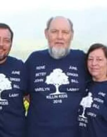 Four people wearing matching navy blue t-shirts with a logo design stand together outdoors. The shirts appear to be for a team or group event.